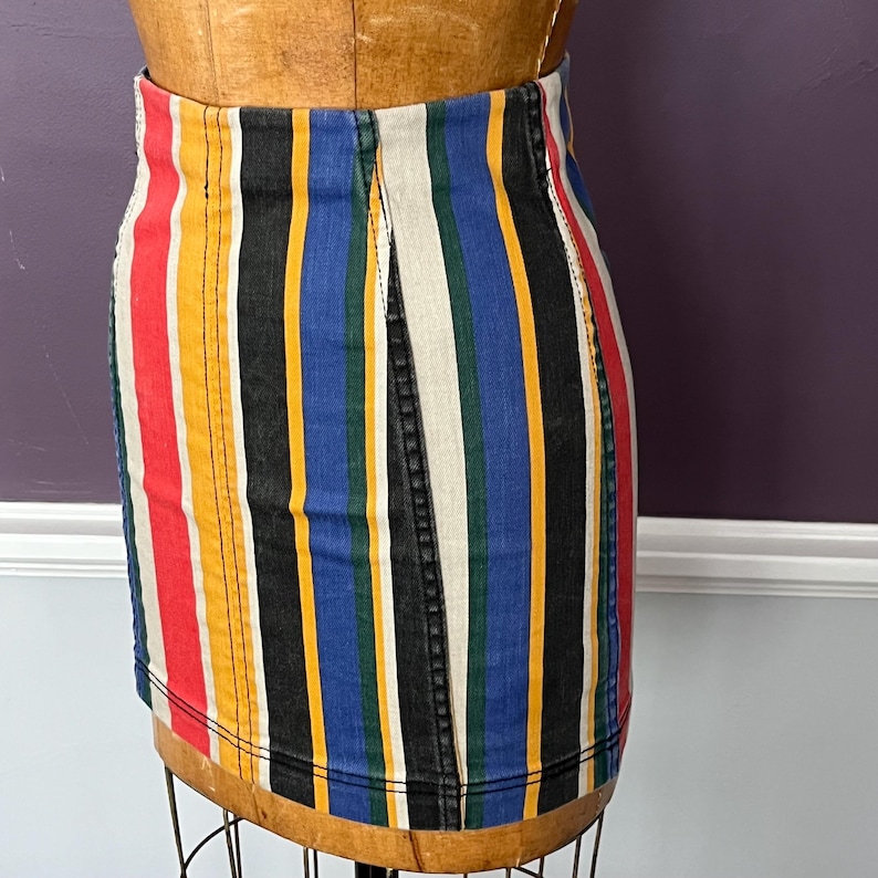 May include: A colorful, striped denim skirt displayed on a mannequin. The skirt features vertical stripes in red, yellow, black, blue, and white. The skirt is a mini-skirt style with a high waist and a classic denim design.