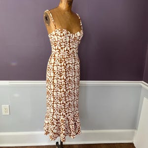 May include: A brown and white floral print dress with spaghetti straps. The dress is displayed on a mannequin. The dress has a fitted bodice and a flared skirt that reaches the ankles. The floral pattern is composed of white flowers on a brown background.