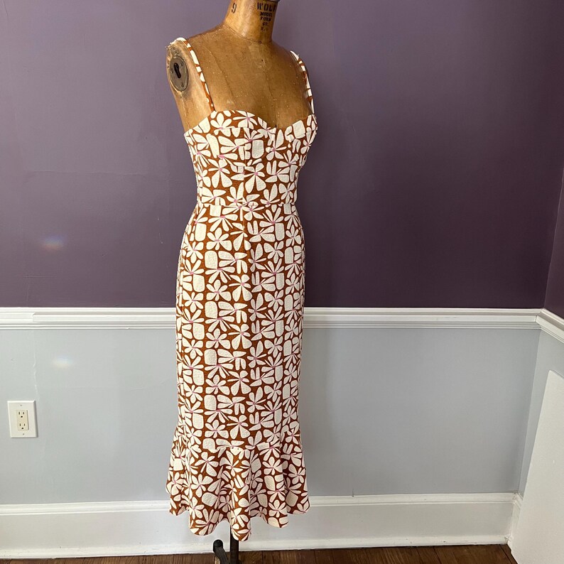 May include: A brown and white floral print dress with spaghetti straps. The dress is displayed on a mannequin. The dress has a fitted bodice and a flared skirt that reaches the ankles. The floral pattern is composed of white flowers on a brown background.