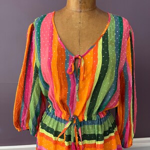 May include: A colorful, striped dress with a V-neckline and long sleeves. The dress features vertical stripes in shades of pink, orange, green, and blue. It has a drawstring waist and a tie at the neckline. The fabric has a dotted texture.