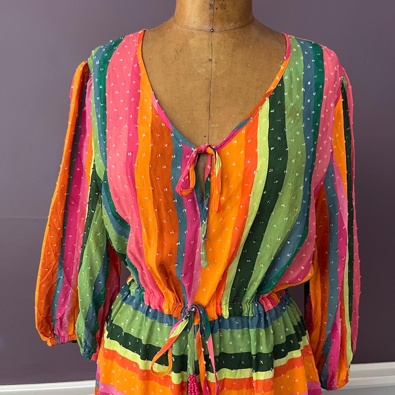 May include: A colorful, striped dress with a V-neckline and long sleeves. The dress features vertical stripes in shades of pink, orange, green, and blue. It has a drawstring waist and a tie at the neckline. The fabric has a dotted texture.