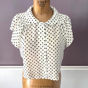 Puede incluir: Una blusa blanca vintage con un estampado de lunares negros. La blusa tiene mangas cortas, cuello redondo y cierre de botones en la parte delantera. La prenda se exhibe en un maniquí vintage.