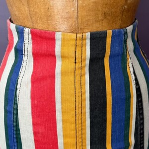 May include: Close-up of a colorful striped skirt. The skirt features vertical stripes in red, white, yellow, black, blue, and green. The fabric appears to be denim or a similar sturdy material, with visible stitching.