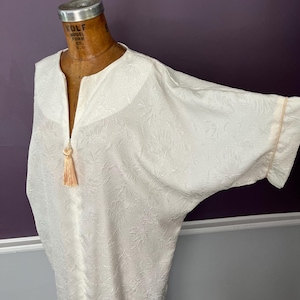 Pode incluir: Vestido de caftan texturizado branco sujo com mangas largas. O vestido apresenta um decote em V com uma borla cor de pêssego. O tecido tem um padrão floral subtil. As mangas têm uma guarnição cor de pêssego.
