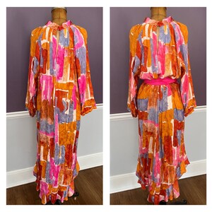 Puede incluir: Un vestido largo y fluido con un vibrante estampado abstracto en tonos naranja, rosa, morado y marr&oacute;n. El vestido presenta mangas largas, un cuello alto y un detalle de ojo de cerradura en el pecho. Un cintur&oacute;n rosa brillante ci&ntilde;e la cintura y el vestido tiene un dobladillo con volantes.