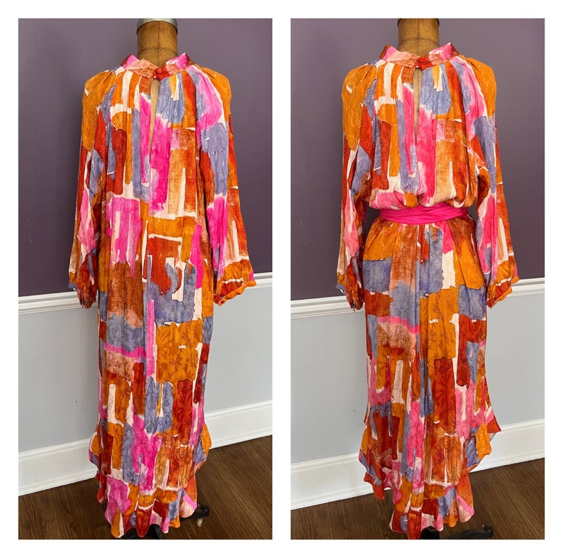 Puede incluir: Un vestido largo y fluido con un vibrante estampado abstracto en tonos naranja, rosa, morado y marr&oacute;n. El vestido presenta mangas largas, un cuello alto y un detalle de ojo de cerradura en el pecho. Un cintur&oacute;n rosa brillante ci&ntilde;e la cintura y el vestido tiene un dobladillo con volantes.
