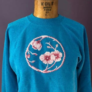 Pode incluir: Uma camisola turquesa com gola redonda e mangas compridas. Apresenta um design floral bordado em ponto cruz em rosa e branco, dentro de uma moldura circular. A camisola é exibida num manequim vintage.
