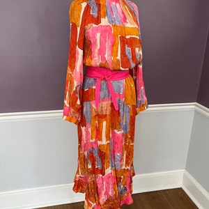 Puede incluir: Un vestido vintage de manga larga, cuello alto y cintur&oacute;n rosa brillante. El vestido presenta un colorido estampado abstracto en tonos naranja, rosa y azul. Tiene un dobladillo con volantes y se exhibe en un maniqu&iacute;.