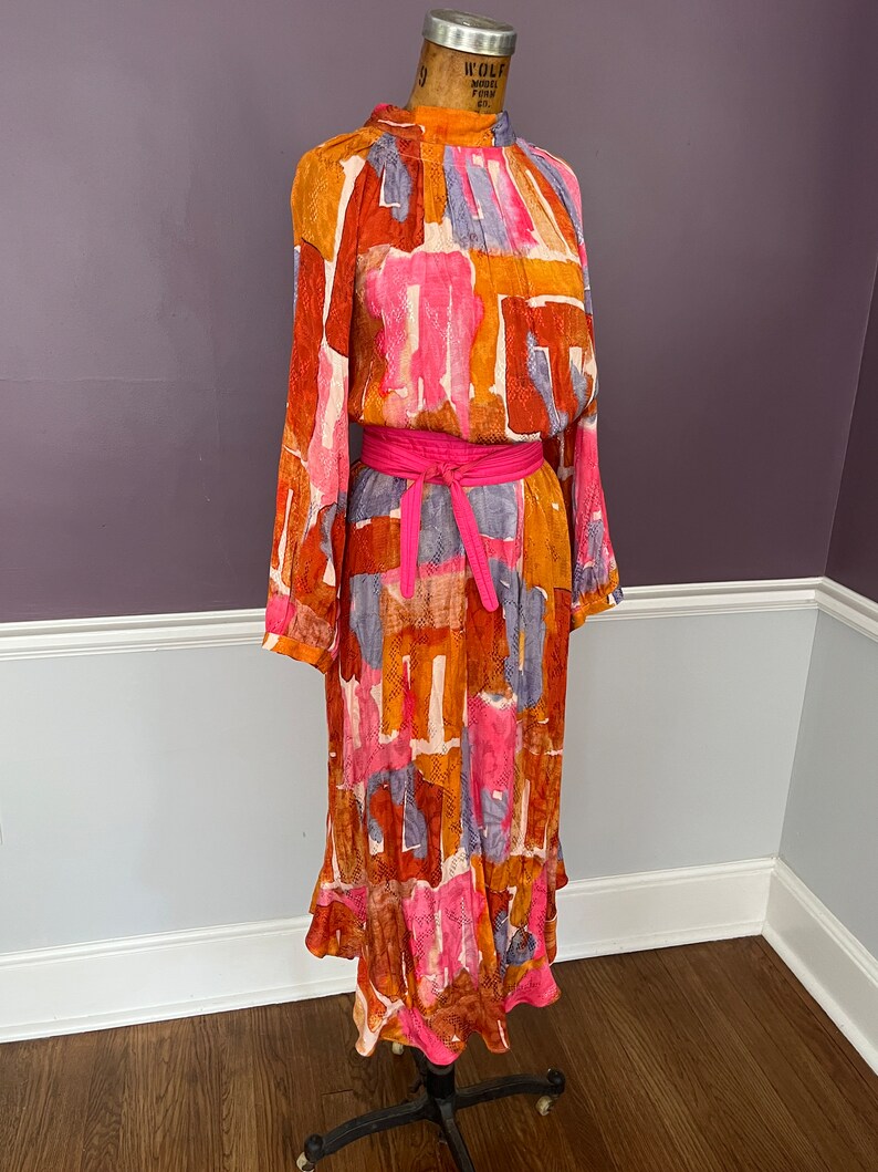 Puede incluir: Un vestido vintage de manga larga, cuello alto y cintur&oacute;n rosa brillante. El vestido presenta un colorido estampado abstracto en tonos naranja, rosa y azul. Tiene un dobladillo con volantes y se exhibe en un maniqu&iacute;.