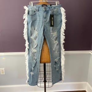 May include: Light wash denim jeans with distressed details and white fringe trim along the outer seams. The jeans have a classic five-pocket design and a black tag. The jeans are displayed on a mannequin.