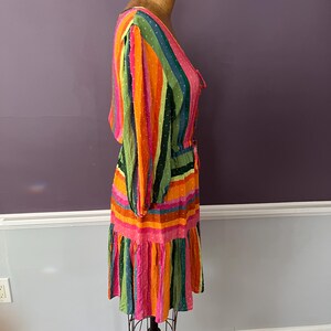 May include: A colorful, striped dress with long sleeves and a ruffled hem. The dress features vertical stripes in shades of pink, orange, green, and blue. The fabric has a dotted texture. The dress is displayed on a mannequin.