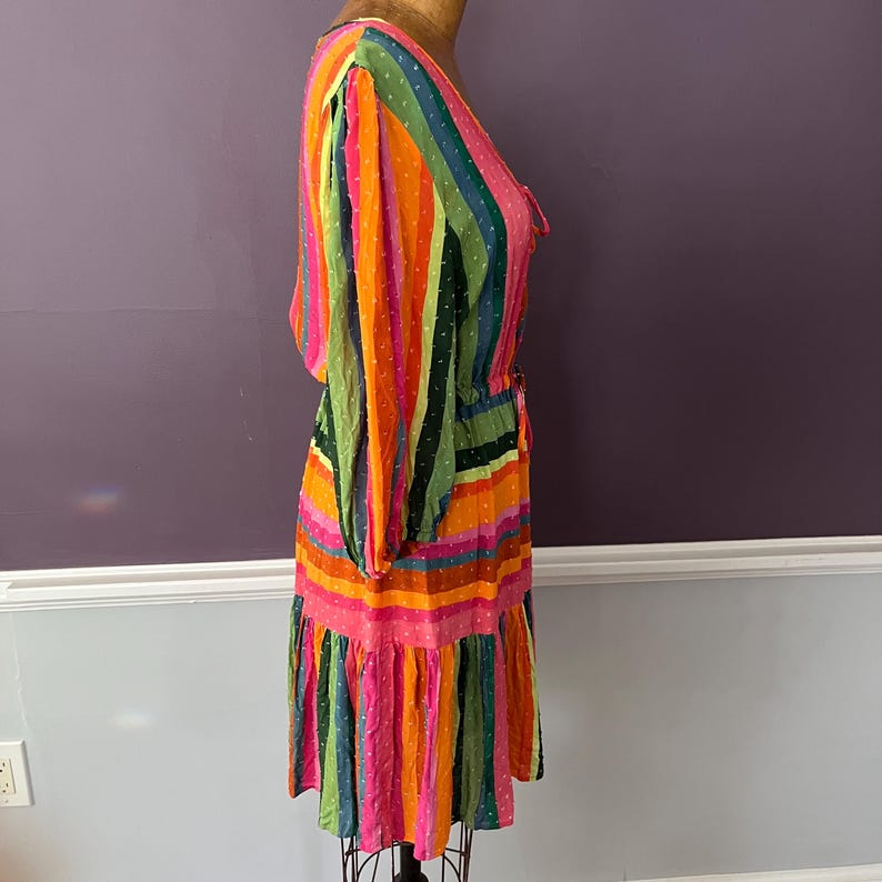 May include: A colorful, striped dress with long sleeves and a ruffled hem. The dress features vertical stripes in shades of pink, orange, green, and blue. The fabric has a dotted texture. The dress is displayed on a mannequin.