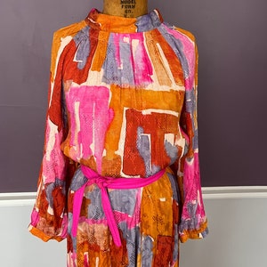 Puede incluir: Un vestido vintage de manga larga, con cuello alto y cintur&oacute;n rosa brillante. El vestido presenta un colorido estampado abstracto en tonos naranja, rosa, marr&oacute;n y gris. Se exhibe en un maniqu&iacute; vintage.