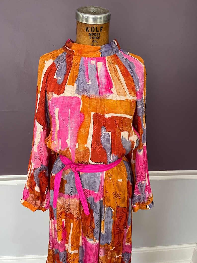 Puede incluir: Un vestido vintage de manga larga, con cuello alto y cintur&oacute;n rosa brillante. El vestido presenta un colorido estampado abstracto en tonos naranja, rosa, marr&oacute;n y gris. Se exhibe en un maniqu&iacute; vintage.