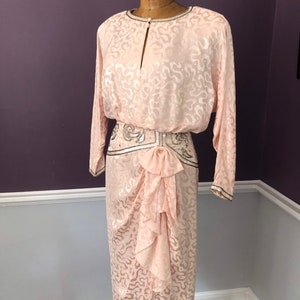 May include: A vintage pale pink dress with a distinctive pattern and long sleeves. The dress has a keyhole neckline, a beaded belt, and a draped skirt with a bow detail. The sleeves and neckline are trimmed with silver beading.