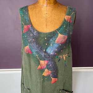 Pode incluir: Vestido sem mangas verde-oliva com decote redondo. O vestido apresenta um design pintado à mão de folhas estilizadas em tons de rosa, laranja e amarelo, com uma libélula e pequenos pontos. O vestido tem dois bolsos.