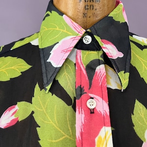 Puede incluir: Primer plano de una camisa negra abotonada con un estampado floral llamativo. El diseño presenta grandes flores rosas, amarillas y blancas con hojas verdes sobre un fondo negro. La camisa está abotonada, mostrando el cuello y los botones.