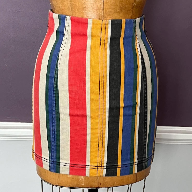 May include: A colorful, vertically striped denim skirt. The skirt features bold stripes of red, cream, yellow, black, blue, and green. The skirt is displayed on a wooden dress form. The skirt has a classic, fitted design.