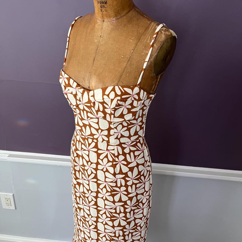 May include: A floral print dress with a brown background and white flower pattern. The dress has thin straps and a fitted bodice. The dress is displayed on a vintage dress form.