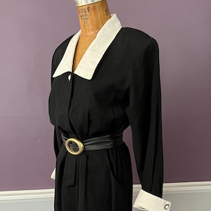 Pode incluir: Vestido preto com gola e punhos brancos, com cinto preto com fivela dourada. O vestido tem mangas compridas e fecho de botões. O vestido é exibido em um manequim.