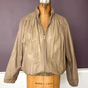 Pode incluir: Casaco bege com gola alta e zíper completo. O casaco tem um design acolchoado nos ombros e é exibido em um manequim vintage. O material do casaco parece ser couro ou um tecido semelhante ao couro.