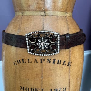 Puede incluir: Un cinturón de cuero marrón con una hebilla decorativa. La hebilla es rectangular con un diseño floral y está incrustada con pequeños diamantes de imitación transparentes. El cinturón está envuelto alrededor de un maniquí vintage. El maniquí tiene la palabra "COLLAPSIBLE".