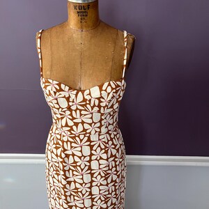 May include: A brown and white floral print dress with thin straps. The dress is displayed on a vintage dress form. The floral pattern features white flowers with pink accents on a brown background. The dress has a fitted bodice and a straight skirt.