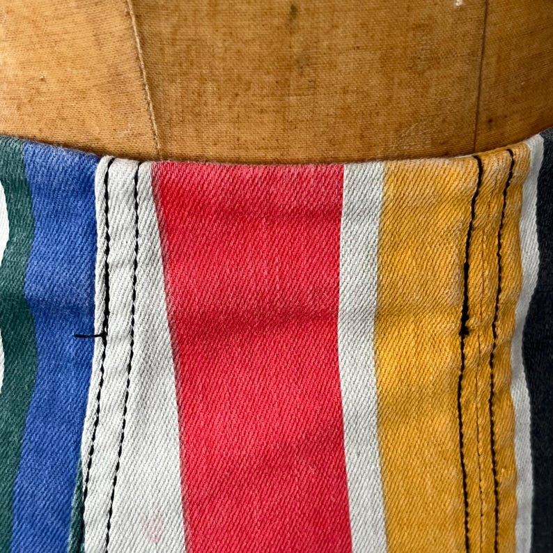 May include: Close-up of a colorful striped fabric, featuring vertical bands of red, yellow, blue, green, and white. The fabric is sewn with black thread, and the texture suggests a denim or canvas material. The item is displayed on a mannequin.