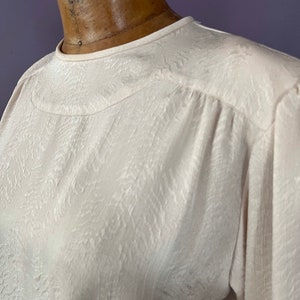 May include: A light cream-colored blouse with a textured, patterned fabric. The blouse has a round neckline and long sleeves. The fabric appears to have a subtle, embossed design. The garment is displayed on a mannequin.