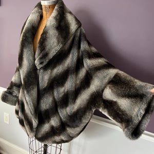 May include: A faux fur coat in shades of grey and black, displayed on a mannequin. The coat features a wide collar and a striped pattern. The sleeves are wide and the coat has a cape-like design.