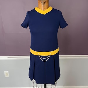 May include: A navy blue short-sleeved dress featuring a yellow V-neck collar and a matching yellow horizontal band at the waist. The dress includes a decorative silver-toned chain draped across the front waistline and a pleated skirt section.