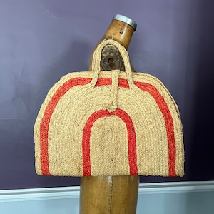 Puede incluir: Un bolso de mano de paja tejida con forma semicircular y dos rayas rojas. El bolso tiene dos asas y un cierre de botón. El bolso se exhibe en un maniquí de madera.