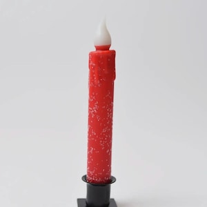 May include: A red candle with a white flame-shaped tip, speckled with white, stands on a black square base. The candle has a dripped wax effect. The candle is a decorative item.