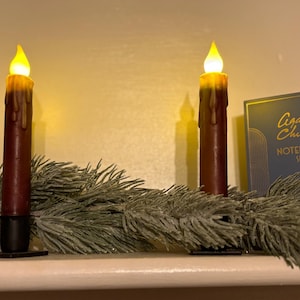 May include: Two battery-operated LED candles with a realistic flickering flame effect. The candles are dark red with a wax-like appearance and are placed on a shelf with a frosted pine garland. A notebook set is visible in the background.
