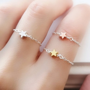 May include: Three delicate star-shaped rings on a finger. The rings are made of silver chain with a small star charm in silver, gold, and rose gold.