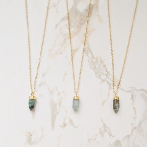 May include: Three gold necklaces with different colored crystal pendants. The pendants are all in the shape of a point. The necklaces are on a white marble background.