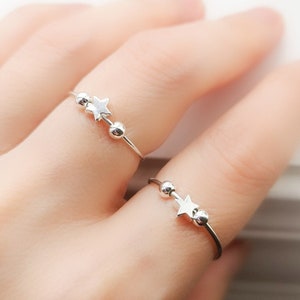 Peut inclure: Deux bagues en argent avec un charme en forme d'étoile et de petites perles argentées. Les bagues sont portées sur le doigt d'une personne.