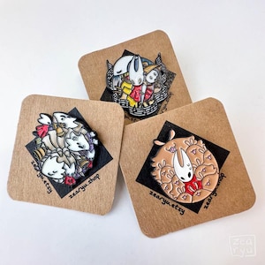 May include: Three enamel pins with cartoon character designs, presented on square cardboard backings. The pins display intricate details and a range of colours, such as white, red, and brown. The text "zearyu.etsy" is printed on the backing.