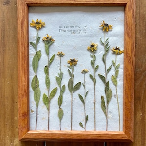 Könnte beinhalten: Ein gerahmtes Kunstwerk mit gepressten gelben Blumen und grünen Blättern auf einem strukturierten weißen Hintergrund. Der Holzrahmen umgibt das Kunstwerk, und der Text "FOR I AM WITH YOU, I WILL TAKE CARE OF YOU. Jeremiah 2:19" ist aufgedruckt.