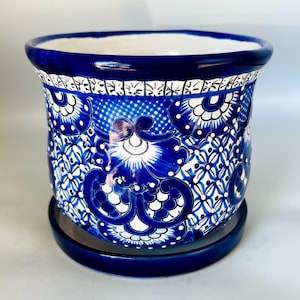 May include: A ceramic planter with a matching saucer, featuring a vibrant blue and white hand-painted floral and geometric pattern. The cylindrical pot has a wide, rounded rim and a glossy finish, showcasing intricate scrollwork and cross-hatching.