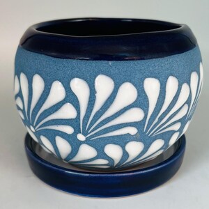 May include: A round ceramic planter with a matte blue textured finish and a glossy dark blue rim. The body features a repeating white floral pattern of teardrop-shaped petals. The planter sits on a matching glossy dark blue circular saucer.