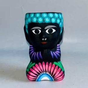 May include: A black ceramic planter shaped like a human figure with a head featuring bright turquoise circular patterns. The figure has painted white eyebrows, black eyes, a red mouth, purple neck accents, and a large pink and green floral design.