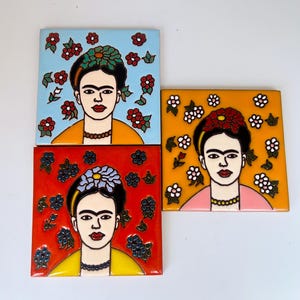 May include: Three square ceramic tiles featuring portraits of Frida Kahlo. One tile has a light blue background with red flowers, one has a bright orange background with white flowers, and one has a red background with blue flowers.