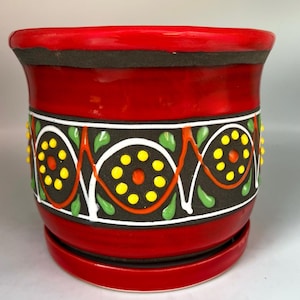 May include: A cylindrical ceramic plant pot with a matching saucer, featuring a glossy red finish on the top and bottom rims. The center band is matte black with a decorative pattern of white swirls, orange zigzags, yellow dots, and green leaves.