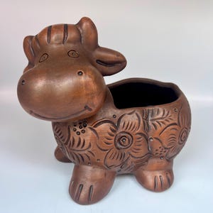 May include: A brown ceramic hippo planter with a large open top. The hippo has a smiling face and floral etched designs on its body. The planter is a decorative item for plants or other small items.