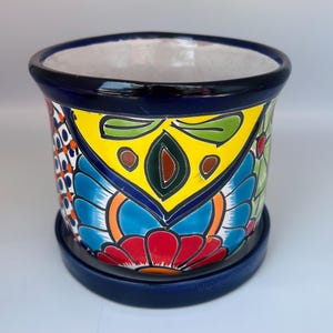May include: A vibrant, hand-painted ceramic flower pot with a matching saucer. The pot features a colorful floral design in yellow, blue, red, and green. The rim and saucer are a deep blue. This decorative pot is ideal for indoor or outdoor plants.