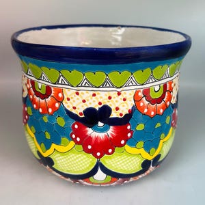 May include: A ceramic planter featuring a vibrant hand-painted pattern with a deep blue rim. The exterior displays a colorful design of red, orange, and lime green floral motifs, intricate geometric shapes, and dotted accents on a white base.