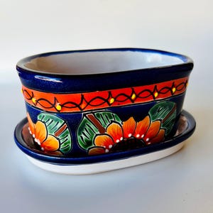 May include: A hand-painted ceramic planter with a matching saucer. The planter is oval-shaped with a deep blue exterior, accented with orange and red floral designs. The interior is white. The saucer is also blue and white, complementing the planter's design.