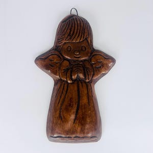 May include: A brown, carved wooden angel figurine with a halo and clasped hands. The angel has detailed wings and a simple, smiling face. A metal loop is attached for hanging.