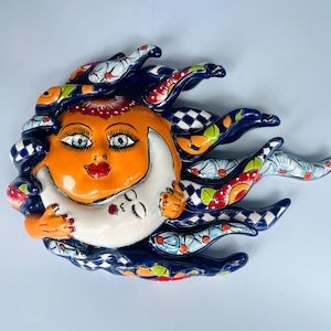 May include: A ceramic wall plaque featuring a bright orange sun face with large eyes and red lips, embracing a white crescent moon face. The design is surrounded by dark blue, flowing rays decorated with checkered patterns, floral motifs, and orange accents.